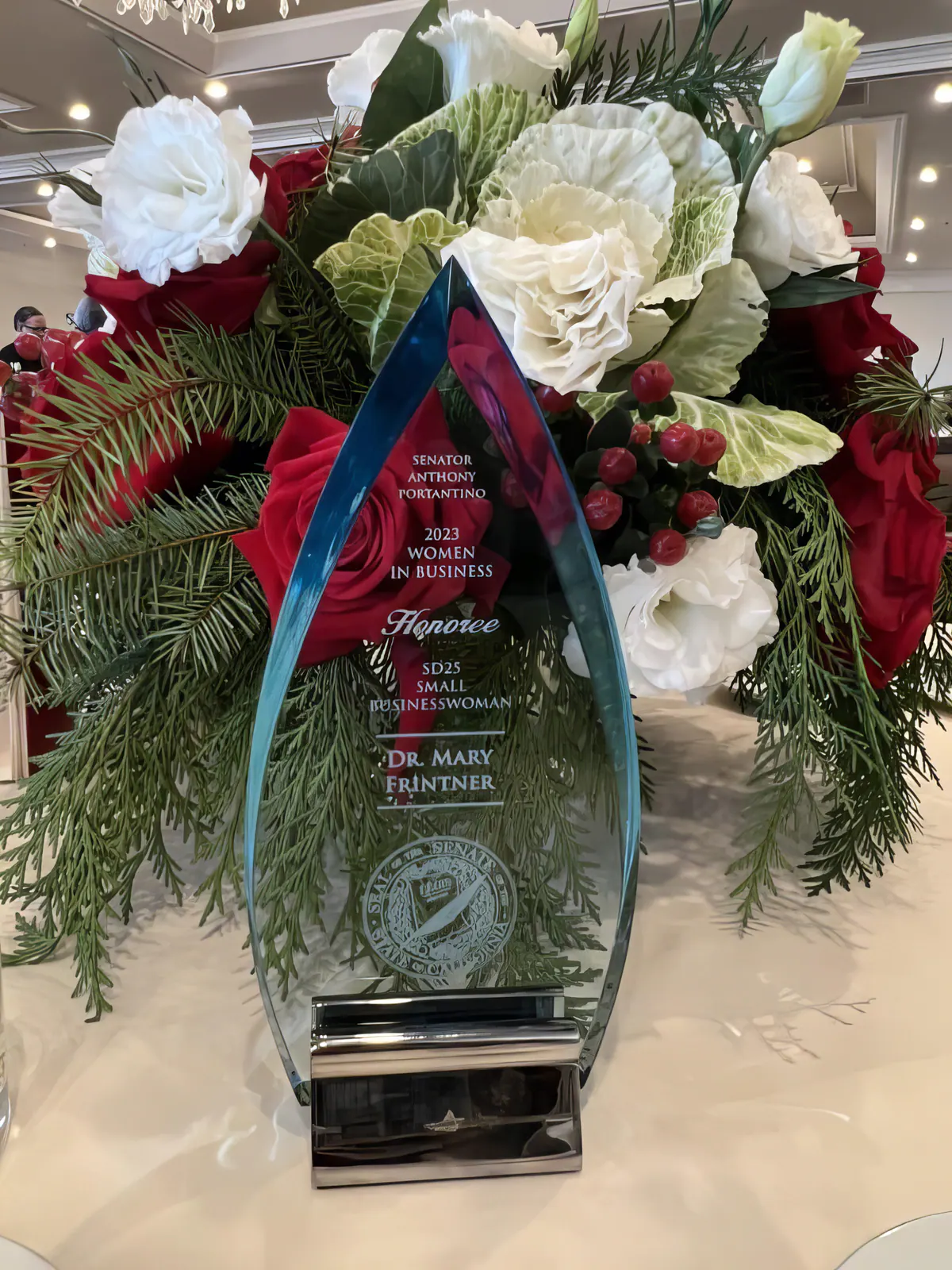 Dr. Mary Frintner’s glass 2023 Women in Business Honoree award for Senate District 25, displayed among red and white flowers